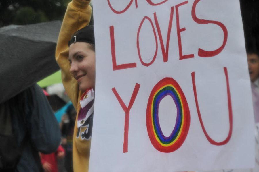 A student wears a rainbow flag tied around her neck and holds a sign that reads God loves you. (Photo by Anna Bongardino | Assistant Visual Editor)